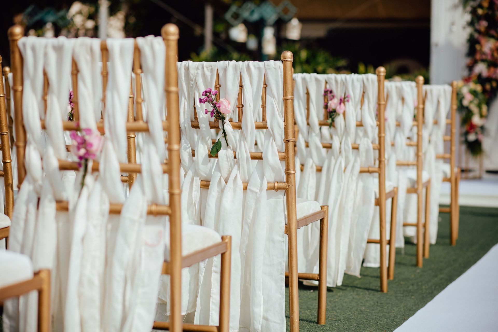 Row of elegant wooden chairs decorated with white fabric and pink flowers for an outdoor wedding ceremony.